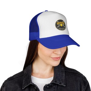 Op de afbeelding: Blauwe en witte truckerpet met een gebogen klep. De pet heeft een patch met de tekst "Honor the Game" en een honkbalafbeelding. De pet heeft een mesh achterkant en een druksluiting.