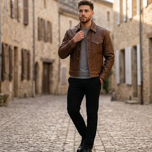May include: A brown leather jacket with a button-down front and two chest pockets. The jacket is worn over a gray shirt and paired with black pants and black shoes. The person is standing on a cobblestone street.