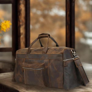 May include: A brown leather duffel bag with a distressed finish. The bag features a top handle, a front zippered pocket, and a detachable shoulder strap. The bag is designed for travel and has a classic, vintage look.
