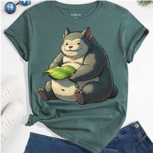 May include: Green t-shirt with a cartoon illustration of a furry creature holding a green leaf. The shirt has rolled-up sleeves and the brand name "SHEIN" is printed on the upper center. The shirt is paired with blue jeans.