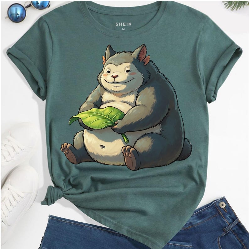 May include: Green t-shirt with a cartoon illustration of a furry creature holding a green leaf. The shirt has rolled-up sleeves and the brand name "SHEIN" is printed on the upper center. The shirt is paired with blue jeans.