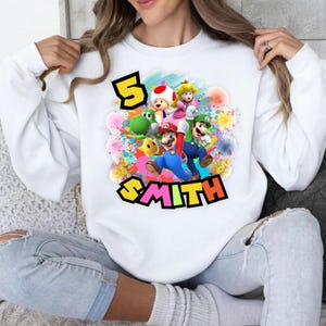 Pu&ograve; includere: Felpa bianca con una grafica colorata di Mario, Luigi, Yoshi e altri personaggi. Il numero "5" e il nome "SMITH" sono visualizzati in un carattere giocoso. Il design &egrave; circondato da schizzi di vernice colorati.