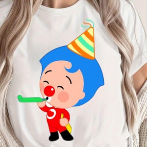 May include: White t-shirt featuring a cartoon character with blue hair, a red nose, and a party hat. The character is wearing a red outfit and blowing a green party horn. The design is centered on the shirt.