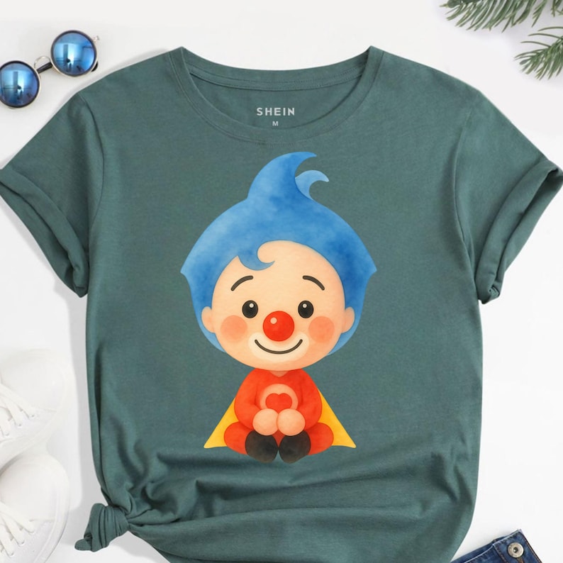 May include: Teal-colored t-shirt featuring a cartoon illustration of a clown with blue hair, a red nose, and a red outfit with a yellow cape. The shirt has rolled-up sleeves and the brand name "SHEIN" is printed on the top.