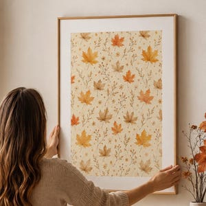 Könnte beinhalten: Ein rechteckiger Wandkunstdruck mit einem sich wiederholenden Muster aus Aquarell-Ahornblättern in Orange-, Gelb- und Brauntönen auf cremefarbenem Hintergrund, eingefasst in einen dünnen, hellen Holzrahmen an einer schlichten Wand.