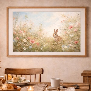 Könnte beinhalten: Ein gerahmtes Aquarellgemälde eines Kaninchens in einem Feld mit Wildblumen hängt über einem Holzesstisch. Der Tisch ist mit Tellern, Tassen und einer Schüssel Brot gedeckt. Eine Lampe und eine Vase mit Trockenblumen stehen auf dem Tisch.