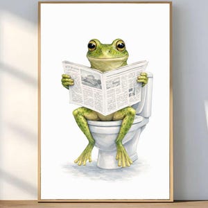 Könnte beinhalten: Gerahmter Kunstdruck, der einen grünen Frosch zeigt, der auf einer weißen Toilette Zeitung liest. Der Frosch hat große Augen und befindet sich in einer humorvollen Pose. Der Druck ist vor einem weißen Hintergrund und in einem hellbraunen Holzrahmen gerahmt.