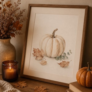 Könnte beinhalten: Ein gerahmter Aquarelldruck mit einem zentralen weißen Kürbis, umgeben von verstreuten braunen Herbstblättern und kleinen grünen Zweigen, alles vor einem neutralen cremefarbenen Hintergrund in einem schlichten Holzrahmen.