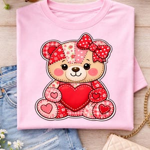 Könnte beinhalten: Rosafarbenes T-Shirt mit einem Patchwork-Teddybär-Design, das ein rotes Herz hält. Der Bär hat eine rote Schleife mit weißen Punkten. Das Design enthält Herzen. Das T-Shirt wird auf einer Holzoberfläche präsentiert.