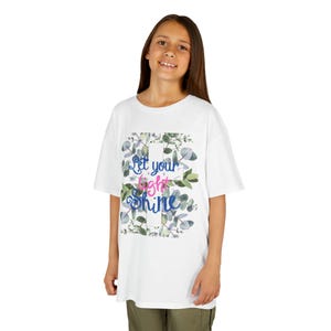 May include: White t-shirt with the text "Let your light shine" in blue and pink script, surrounded by a floral design with green and blue leaves. The shirt is worn by a person.