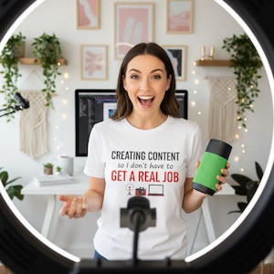 May include: A white t-shirt featuring the black and red text "CREATING CONTENT so I don't have to GET A REAL JOB" with small icons of a ring light, a camera, and a laptop. The shirt is worn by a person holding a green and black cylindrical speaker.