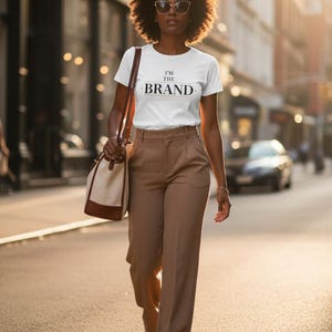 May include: A white short-sleeved t-shirt with the text I'M THE BRAND printed in black on the chest, paired with high-waisted tan trousers, white sneakers, and a two-toned canvas and brown leather shoulder bag with thin brown straps.