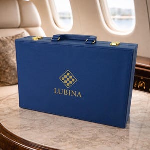 Może przedstawiać: Niebieska skórzana teczka ze złotymi okuciami, uchwytem i zapięciami. Teczka ma słowo "LUBINA" wydrukowane w kolorze złotym pod logo w kształcie rombu z wzorem szachownicy. Teczka leży na marmurowym stole.