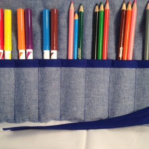 Puede incluir: Un estuche enrollable de mezclilla azul con cierre de cinta azul contiene una variedad de marcadores y lápices de colores.