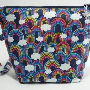 May include: A navy blue zipped pouch featuring a repeating pattern of colourful rainbows and white clouds. The pouch includes a matching fabric wrist strap with a silver-toned metal clasp and a small silver sheep-shaped charm on the zip pull.