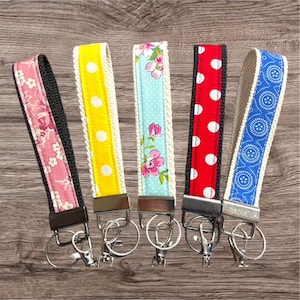 May include: Five key fobs with diverse fabric patterns. The patterns include pink floral, yellow with white polka dots, light blue with pink flowers, red with white polka dots, and blue with a white circular design. Each fob has a metal clasp and key ring.