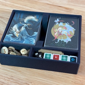 May include: A navy blue game organizer with compartments for cards, tokens, and dice. The cards feature fantasy art with blue and gold accents. The organizer also holds wooden tokens and small, colorful game pieces.