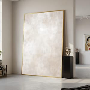 May include: A large abstract painting with a gold frame leans against a white wall. The artwork features a soft, textured design in shades of beige and cream. The room includes black pedestals with sculptures and another framed artwork.