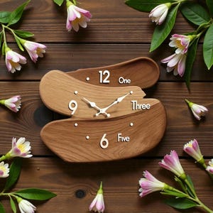 Op de afbeelding: Een houten wandklok met een uniek, gelaagd ontwerp. De klok heeft een lichtbruine houtnerf met witte cijfers op 12, 9, 6 en 3 uur, en de woorden "One", "Three" en "Five". De wijzers zijn zilverkleurig. De klok is omringd door bloemen.