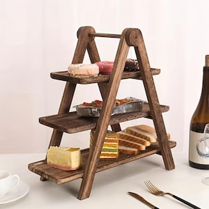 May include: A three-tiered, wooden serving stand with a ladder-like design. The stand holds various pastries, including donuts, cake, and sandwiches. The wood has a dark brown finish. A bottle of wine and a fork and knife are also visible.