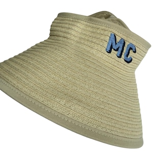 May include: A beige straw sun visor featuring a wide, structured brim and a matching headband. The front of the visor is embroidered with the letters MC in light blue thread with a dark blue outline, providing a clear contrast against the straw.
