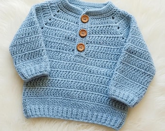 May include: A light blue crochet sweater with a round neckline and long sleeves. The sweater features three wooden buttons down the front. The crochet stitch creates a textured, horizontal striped pattern. A cozy, handmade garment.