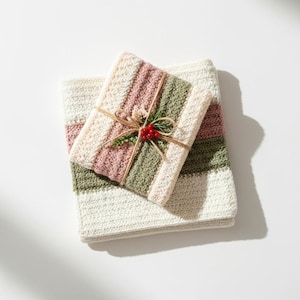 May include: A folded, striped crochet blanket with a smaller, wrapped crochet item on top. The blanket and wrapped item feature stripes of cream, dusty rose, and olive green. The wrapped item is tied with twine and decorated with a sprig and red berries.