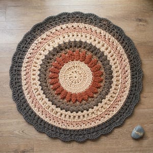 May include: A round, handmade crochet rug with a scalloped edge. The rug features concentric circles in shades of beige, pink, brown, and gray. The center has a sunflower-like design in beige and rust-orange. The rug is placed on a wooden floor.
