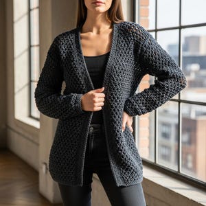 以下が含まれることがあります： テクスチャード加工されたかぎ針編みのデザインが施された、ダークグレーのオープンフロントカーディガン。長袖セーターは膝丈で、太い糸でできているようです。カーディガンは黒いトップスと濃い色のジーンズの上に着用されています。