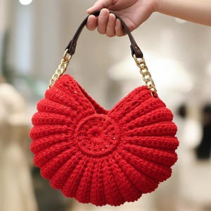 May include: A vibrant red, handmade crochet handbag with a distinctive, fan-shaped design. The bag has a brown leather handle with gold chain detailing. The central design is circular, and the bag is held by a hand.