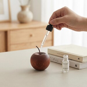 Peut inclure: Un diffuseur en bois brun foncé en forme de pomme avec une tige, une pipette en verre transparent avec un bouchon noir et un petit flacon en verre transparent étiqueté JAM MOON #004 ONE DAY. La pipette est tenue au-dessus de la pomme en bois.