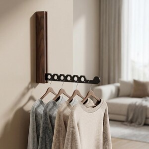 May include: A wall-mounted, folding garment rack made of dark brown wood. The rack features a vertical base attached to the wall and a horizontal arm with six circular cutouts for hangers, currently holding four sweaters on wooden hangers.