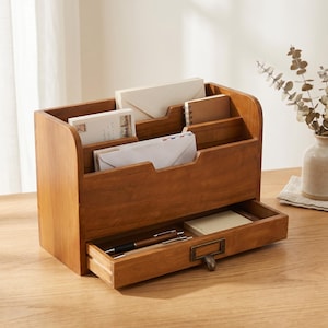 May include: A medium-toned wooden desk organiser featuring multiple vertical slots for mail, envelopes, and notebooks. The unit includes a single pull-out drawer at the base with a brass-coloured metal label holder and handle, containing two pens.