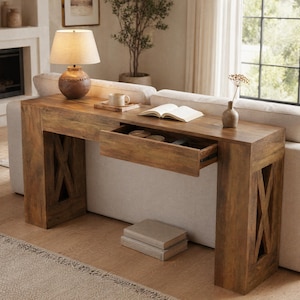 May include: A rectangular wooden console table with a medium brown finish, featuring a single pull-out drawer, X-shaped side supports, and a flat top surface. The table is positioned behind a sofa and holds a lamp, a mug, and a book.