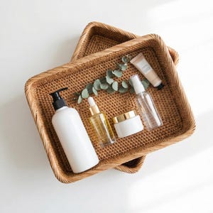 Puede incluir: Dos cestas rectangulares de ratán trenzado encajadas, que contienen una botella con dosificador blanca, un frasco cuentagotas de cristal transparente con detalles dorados, un tarro blanco con tapa dorada, un pulverizador, un tubo beige y una rama de eucalipto.