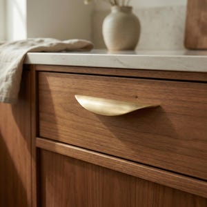 Op de afbeelding: Een gebogen ladehandgreep van geborsteld messing gemonteerd op een houten lade in een middelbruine tint. De handgreep heeft een gladde, halvemaanvormige vorm met een metalen afwerking die contrasteert met de natuurlijke houtnerf.