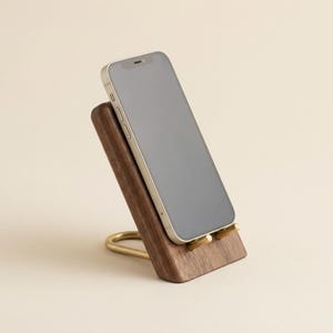Può includere: Uno smartphone appoggiato su un supporto in legno marrone scuro con una base in metallo dorato. Il supporto presenta un blocco di legno inclinato per sostenere il dispositivo e due piccoli pioli dorati sulla parte inferiore.