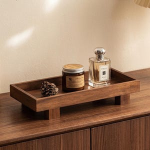Könnte beinhalten: Ein rechteckiges Holztablett mit vier kurzen Füßen, auf dem sich ein braunes Kerzenglas mit dem Etikett Aroma Sensations, ein durchsichtiger Glasparfümflakon mit silbernem Deckel und weißem Etikett sowie ein kleiner brauner Kiefernzapfen befinden.