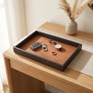 Può includere: Un vassoio rettangolare con una cornice in legno marrone scuro e una base in pelle intrecciata color cuoio. Il vassoio contiene una chiave dell'auto argentata con portachiavi in metallo, un orologio con cinturino in pelle marrone e un anello dorato.
