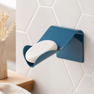 Può includere: Un portasapone da parete blu scuro con un design curvo a forma di scivolo. Una saponetta bianca di forma ovale poggia saldamente al centro del supporto, che è fissato su una superficie piastrellata bianca con un motivo esagonale.