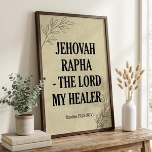 Puede incluir: Arte mural enmarcado con el texto "JEHOVAH RAPHA - THE LORD MY HEALER" en negro sobre un fondo beige. La obra de arte está en un marco marrón oscuro y se exhibe sobre una mesa de madera con plantas decorativas.
