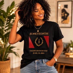 May include: Black t-shirt featuring the word "Juneteenth" above a graphic flag design with red and black sections, a gold wreath, and a sword. The text "Emancipation Day June 19, 1865" is below. The shirt is worn by a person.
