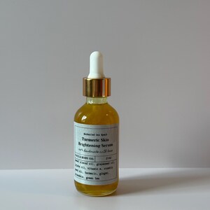 May include: A clear glass bottle filled with a yellow liquid, labeled "Turmeric Skin Brightening Serum." The bottle has a white dropper top with a gold band and a white label with black text. The product is a face and body oil.