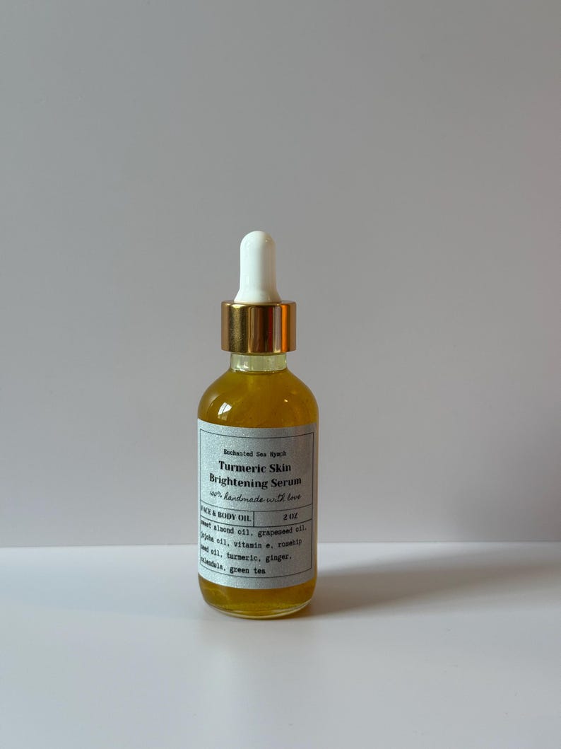 May include: A clear glass bottle filled with a yellow liquid, labeled "Turmeric Skin Brightening Serum." The bottle has a white dropper top with a gold band and a white label with black text. The product is a face and body oil.