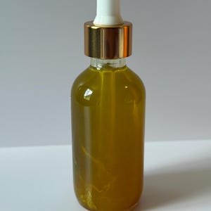 May include: A clear glass bottle filled with a yellow liquid. The bottle has a gold-colored cap and a white dropper. The bottle is cylindrical and sits on a white surface.