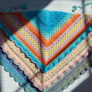 May include: A stack of three triangular crochet shawls featuring scalloped edges and tie strings at the corners. The top shawl displays a vibrant gradient of turquoise, orange, and yellow. The middle shawl is pastel pink and cream, while the bottom shawl is dark blue and purple.
