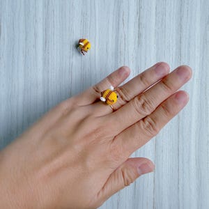May include: A handmade crochet ring featuring a yellow and brown bee design. The bee has white wings and is set on a gold-colored ring band. A second bee is visible in the background. The ring is a unique, whimsical accessory.