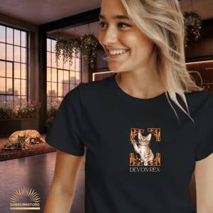 Könnte beinhalten: Ein schwarzes T-Shirt mit einem Grafikdruck einer braunen getigerten Devon Rex Katze, zentriert in einem orange-schwarzen dekorativen Rahmen. Unter der Katzenillustration steht der Text DEVON REX in einer weißen, serifenlosen Schrift.