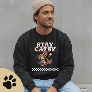 Könnte beinhalten: Ein schwarzes Sweatshirt mit Rundhalsausschnitt und dem Schriftzug STAY CATSY in weißen und roten Buchstaben über einer Cartoon-Katze mit roter Kappe auf einem Skateboard. Unter der Katze befindet sich ein weiß-schwarzes Karomuster.