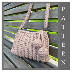 Puede incluir: Un bolso de ganchillo rosa claro con una correa de hombro trenzada gruesa, un cuerpo rectangular con textura acanalada vertical y dos pompones esféricos colgantes unidos a un anillo circular oscuro. La palabra PATTERN está impresa verticalmente.